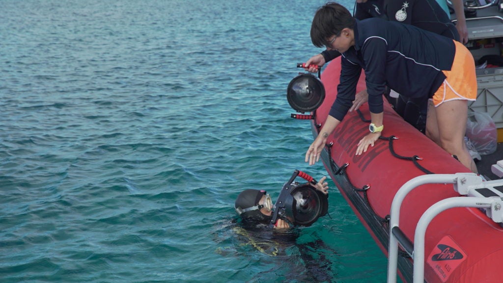 A diver hands a camera to a colleague onboard a research boat